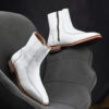 Pair of handcrafted white leather boots with intricate stitching, wooden sole, and side zipper.